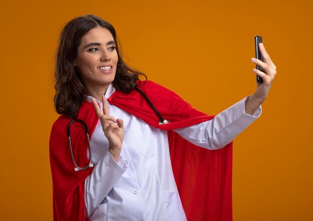 Smiling caucasian superhero girl in doctor uniform with red cape and stethoscope gestures victory hand sign