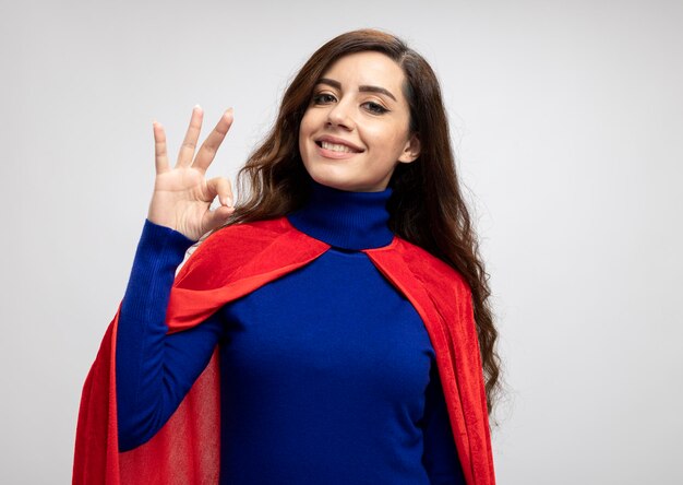 Улыбающаяся кавказская девушка-супергерой с красной накидкой жестами показывает знак рукой на белом