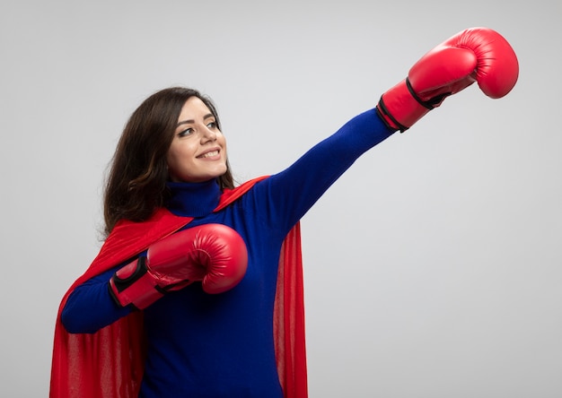 Free photo smiling caucasian superhero girl with red cape wearing wearing boxing gloves stands with raised hands looking and pointing at side isolated on white wall with copy space