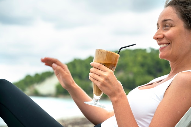 Улыбающаяся кавказская женщина держит кофейный напиток на пляже с пеной и соломинкой с холмами