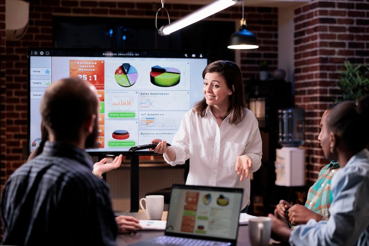 Free Photo smiling caucasian woman in startup office doing business presentation on big tv screen with charts in front of team. confident employee presenting growing sales statistics in late night meeting.
