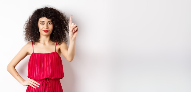 Foto gratuita sorridente donna caucasica con capelli ricci che indossa un abito rosso che mostra il gesto della regola numero uno alzando fi