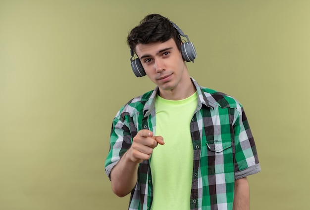 Smiling caucasian young guy wearing green shirt in headphones showing you gesture on isolated green background