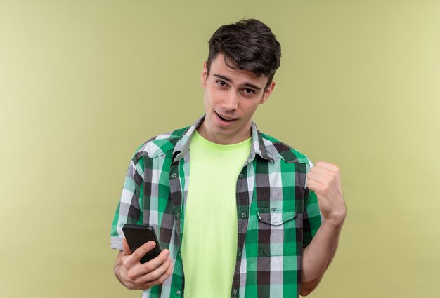Smiling caucasian young guy wearing green shirt holding phone showing yes gesture on isolated green background