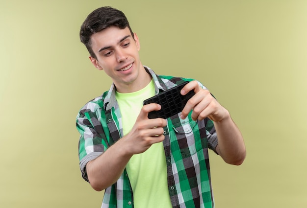 Ragazzo giovane caucasico sorridente che indossa la camicia verde che gioca gioco sul telefono su fondo verde isolato