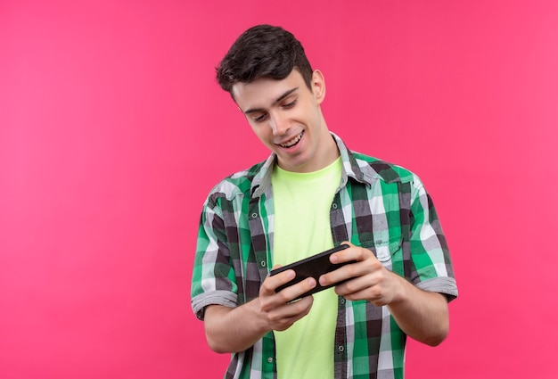 Smiling caucasian young guy wearing green shirt playing game on phone on isolated pink background
