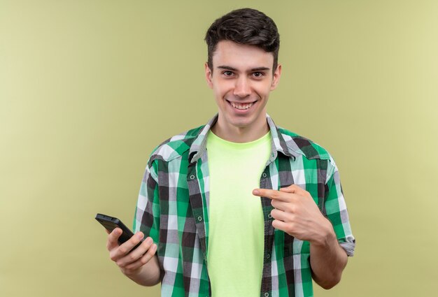 Smiling caucasian young guy wearing green shirt points to phone in his hand on isolated green background