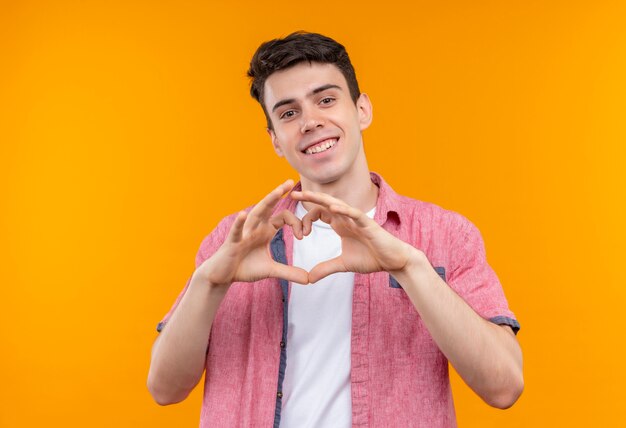 Smiling caucasian young guy wearing pink shirt showing heart gesture on isolated orange background