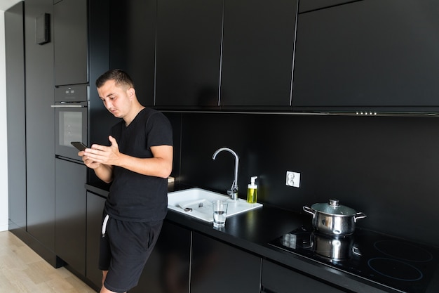 Улыбающийся кавказский молодой человек использует мобильный телефон гаджет для обмена текстовыми сообщениями, стоя на кухне