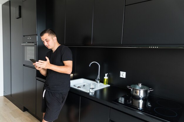 Smiling Caucasian young man use cellphone gadget texting messaging standing in kitchen