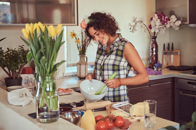웃고 있는 매력적인 곱슬머리 히스패닉계 소녀는 부엌에서 요리하는 동안 재료를 섞습니다.