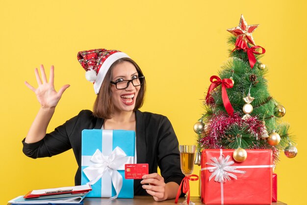 Smiling charming lady in suit with santa claus hat and eyeglasses showing five and holding gift and bank card in the office on yellow isolated 