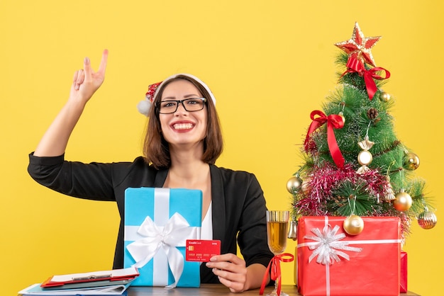 Smiling charming lady in suit with santa claus hat and eyeglasses showing gift and bank card pointing up in the office on yellow isolated 