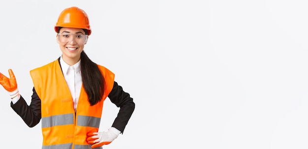 Smiling cheerful asian female construction manager introduce her interprise for investors wearing safety glasses helmet and gloves pointing hand left and looking proud white background