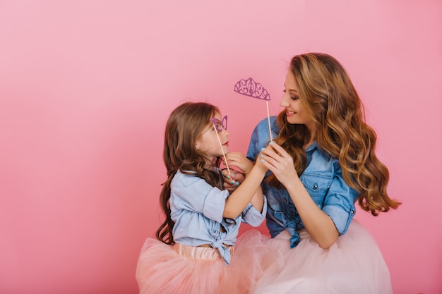 Smiling cheerful young mother looking with love at her long-haired daughter wearing purple carnival mask. Adorable little girl in denim shirt having fun and playing with mom, holding her hands