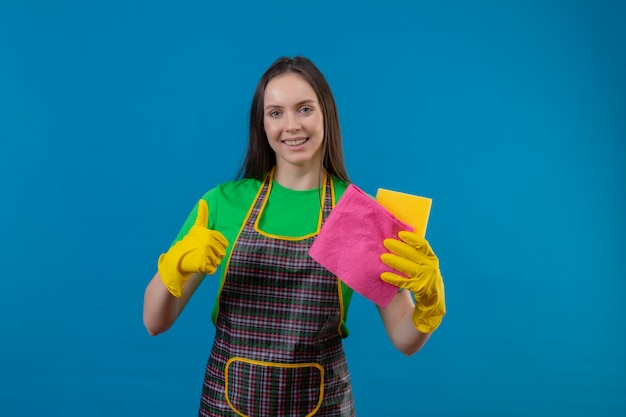 Smiling cleaning young girl wearing uniform in gloves holding cleaning tools her thumb up on isolated blue background