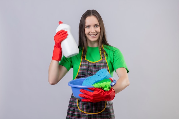 Smiling cleaning young girl wearing uniform in red gloves holding cleaning tools and cleaning agent on isolated white background