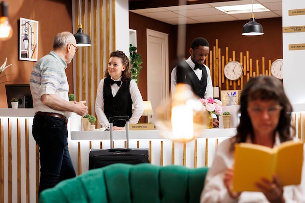 Foto gratuita il concierge sorridente assiste gli ospiti anziani di sesso maschile alla reception, mentre una donna anziana caucasica si diverte a leggere un romanzo nell'area lounge. la lobby dell'hotel dispone di un comodo divano e di un bancone della reception per le prenotazioni.