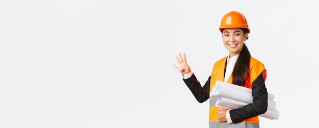 Smiling confident asian female engineer construction manager in safety helmet carry blueprints showing four fingers ensure building finished in time standing white background upbeat