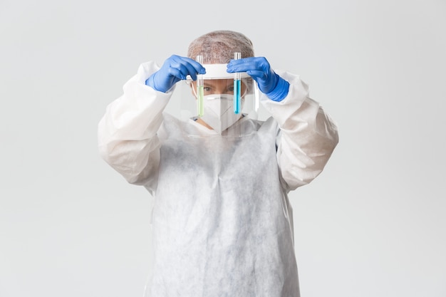 Smiling confident female doctor in personal protective equipment showing two test-tubes with vaccines or medication, 