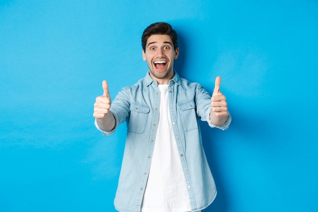 Smiling confident man showing thumbs up with excited face, like something awesome, approving product, standing against blue background