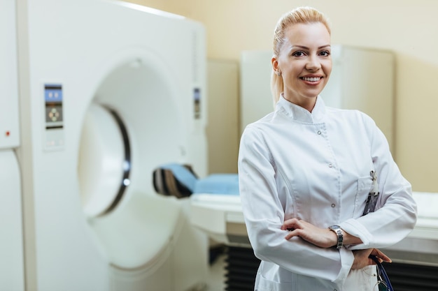 Sorridente radiologo fiducioso in piedi con le braccia incrociate nella stanza dell'esame medico e guardando la fotocamera