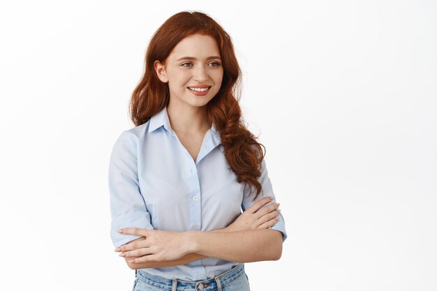 Smiling confident redhead female entrepreneur, looking aside at client, cross arms on chest and looking friendly, chatting, standing casually relaxed against white background