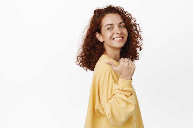 Smiling confident woman with red curly hairstyle, pointing finger behind her, showing smth left, standing over white background