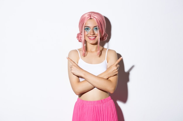 Улыбающаяся уверенная в себе молодая женщина в праздничном костюме и розовом парике выглядит довольной, указывая пальцами в сторону
