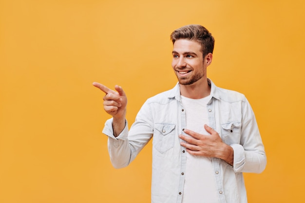 Smiling cool young man with red beard and brown hair in stylish white clothes looking away and showing aside his finger