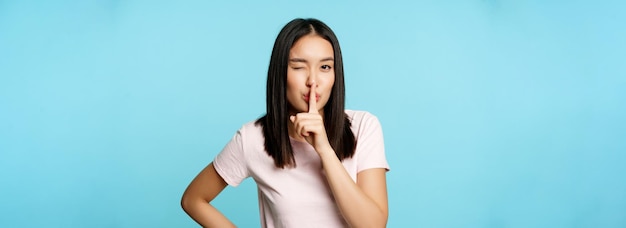Улыбающаяся кокетливая азиатская девушка шикает, заставляя молчать жестом табу, подмигивая и делая знак тишины перед