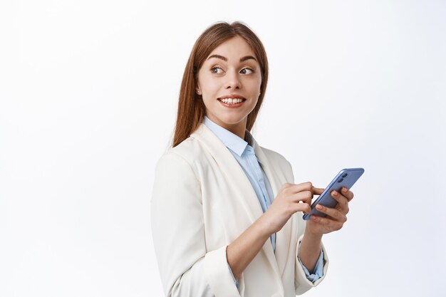 Smiling corporate girl in suit using mobile phone, turn head behind and look at promotional text, reading copy space on white wall, holding smartphone