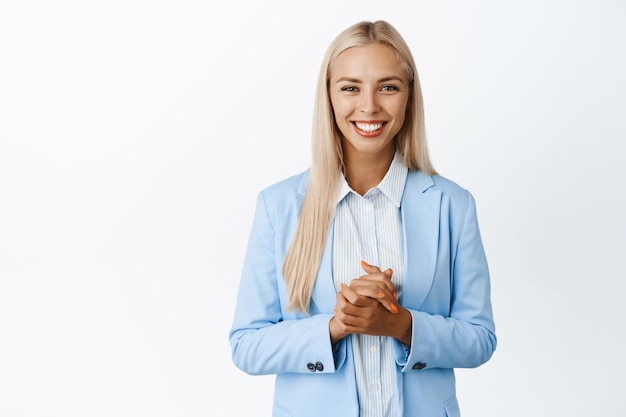 Smiling corporate woman holding hands together looking at client standing in saleswoman suit against white background represent her company