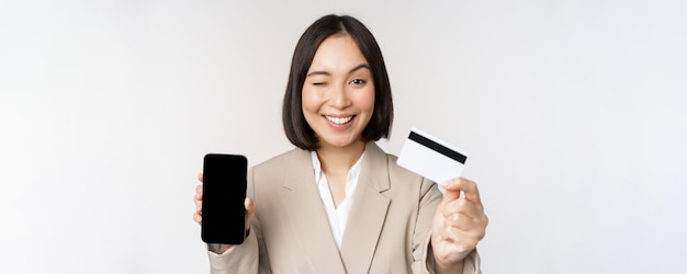 흰색 배경 위에 서 있는 휴대전화 스마트폰 화면에 휴대전화 화면과 앱을 보여주는 정장을 입은 웃고 있는 기업 여성