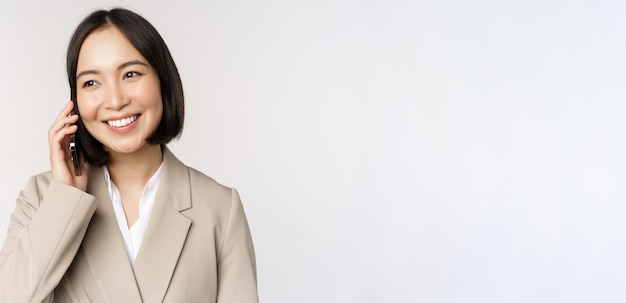 Smiling corporate woman in suit talking on mobile phone having a business call on smartphone standing over white background