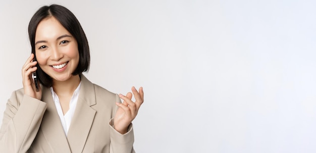 Smiling corporate woman in suit talking on mobile phone having a business call on smartphone standing over white background