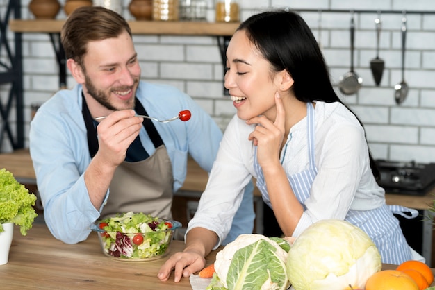 Smiling couple feeding healthy salad in kitchen wearing apron
