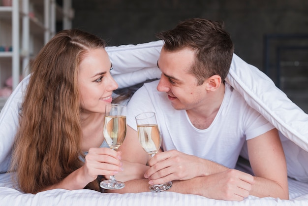 Smiling couple lying on bed under duvet with glasses of drink
