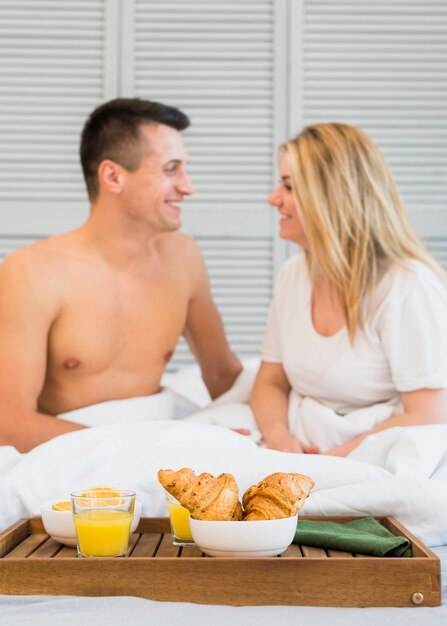 Smiling couple sitting on bed near food on breakfast table 