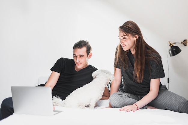 Smiling couple sitting with dog on cozy bed looking at laptop