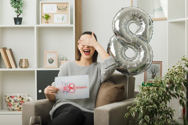 Smiling covered eyes with hand beautiful girl on happy women day holding greeting card sitting on armchair in living room