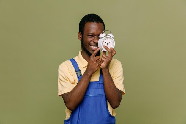 Smiling covered face with alarm clock young africanamerican cleaner male in uniform with gloves isolated on green background