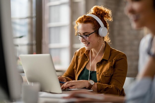 Smiling creative businesswoman listening music on headphones while working on a computer in the office