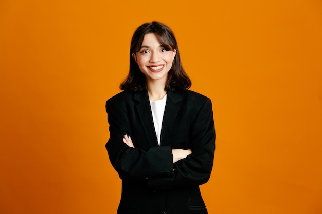 Smiling crossing hands young beautiful female wearing black jacket isolated on orange background