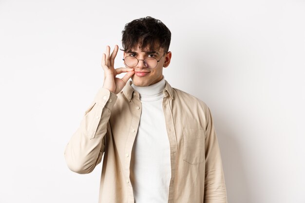 Smiling cunning guy in glasses shut his mouth on zipper, showing seal gesture near lips and smiling devious, keeping a secret, standing on white background