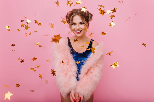 Smiling curly girl in trendy coat arranged a holiday surprise for friend's birthday party. Laughing amazing young woman gladly posing with golden glitter confetti standing on pink background