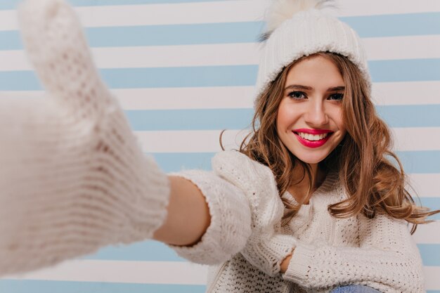 Smiling, curly Slavic model with gentle make-up taking selfie. Portrait of girl in knitted clothes on striped blue wall