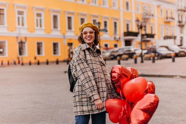 Улыбающаяся кудрявая женщина в оранжевых очках смотрит вперед напротив стены красивого здания