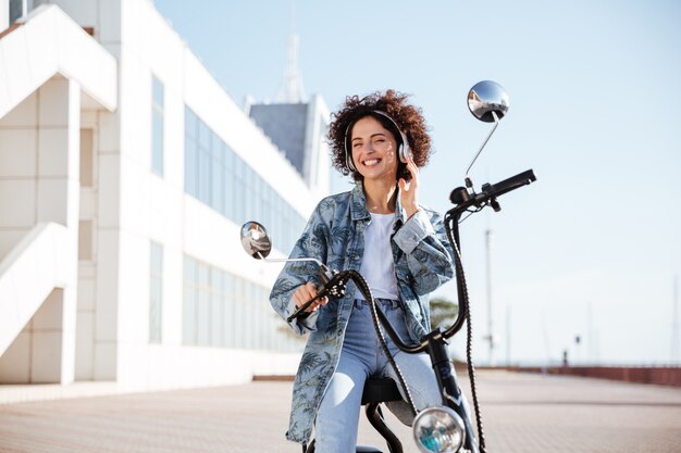 Улыбающаяся кудрявая женщина сидит на современном мотоцикле на свежем воздухе и слушает музыку