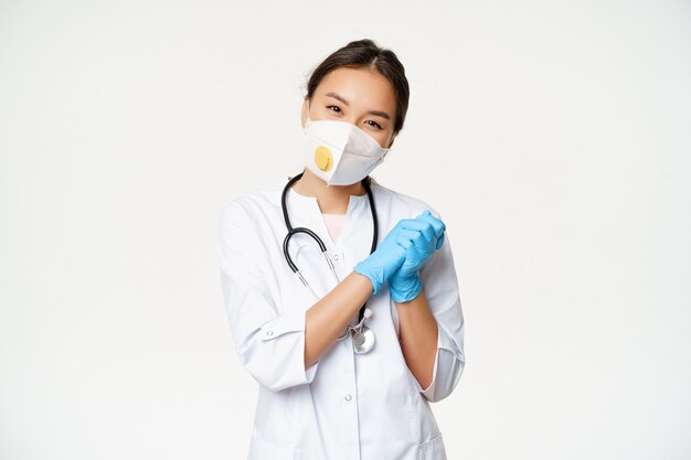 Smiling cute asian female doctor nurse in medical respirator and rubber sterile gloves looking pleas...